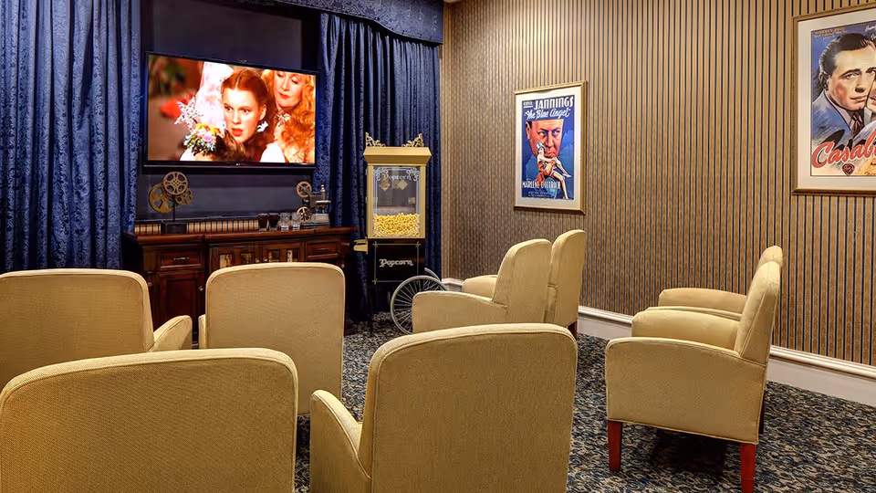 A cozy movie room with six beige armchairs arranged in two rows facing a flat-screen TV mounted on a wall with dark blue curtains. Below the TV is a wooden cabinet with vintage film reels and a popcorn machine filled with popcorn. The walls have striped wallpaper and framed classic movie posters.
