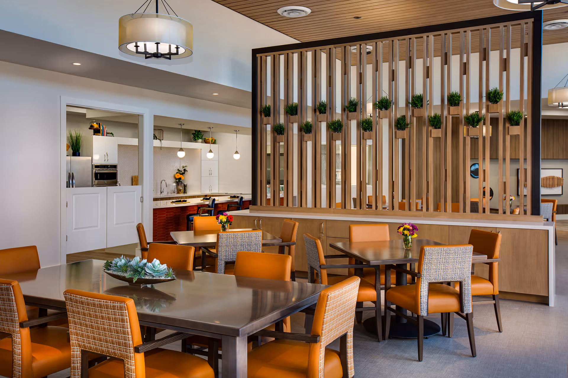 A modern dining area with several tables and orange cushioned chairs. A wooden partition with small green plants separates the dining space from an adjacent kitchen area, which features white cabinets, pendant lights, and stainless steel appliances. The room has a light and airy atmosphere with a contemporary ceiling light fixture.