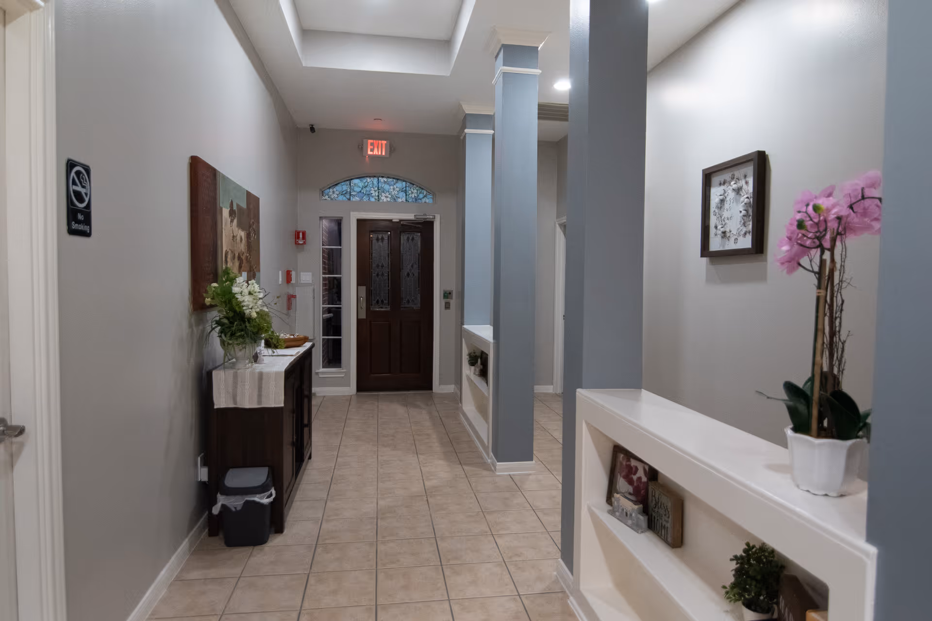 A well-lit hallway with tiled floor and light gray walls. On the left side, there is a small wooden table with a white runner, a vase with white flowers, and a no smoking sign on the wall. On the right side, there are built-in shelves with decorative items and a potted pink orchid on top. At the end of the hallway, there is a dark wooden door with glass panels and an exit sign above it.