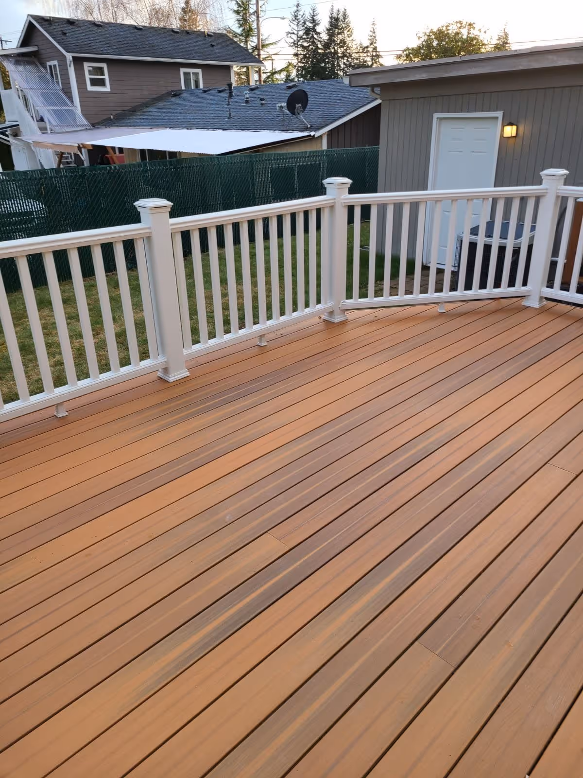 A wooden outdoor deck with light brown planks and white railing. In the background, there is a green chain-link fence, a gray building with a white door and a light fixture, and neighboring houses with trees behind them.