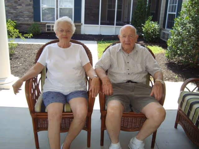An elderly woman and an elderly man sitting side by side on wicker chairs on a covered patio outside a residential building, with plants and windows visible in the background.