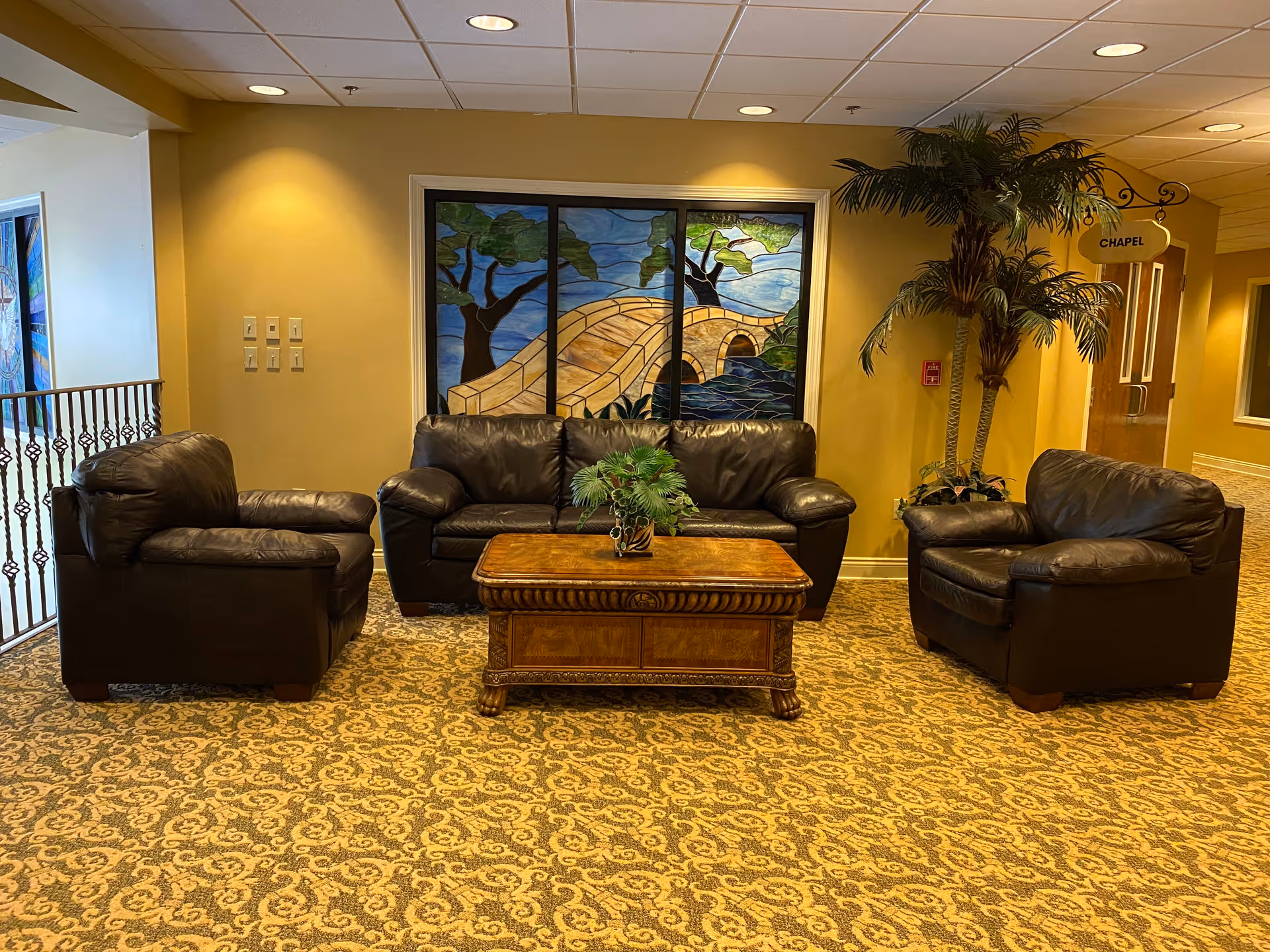A cozy seating area in a senior living facility with a dark leather sofa and two matching armchairs arranged around a wooden coffee table with a small plant on it. Behind the sofa is a large stained glass window depicting a bridge and trees. To the right, there is a tall potted plant and a hallway with a sign labeled 'Chapel'. The walls are painted yellow and the floor is covered with a patterned carpet.