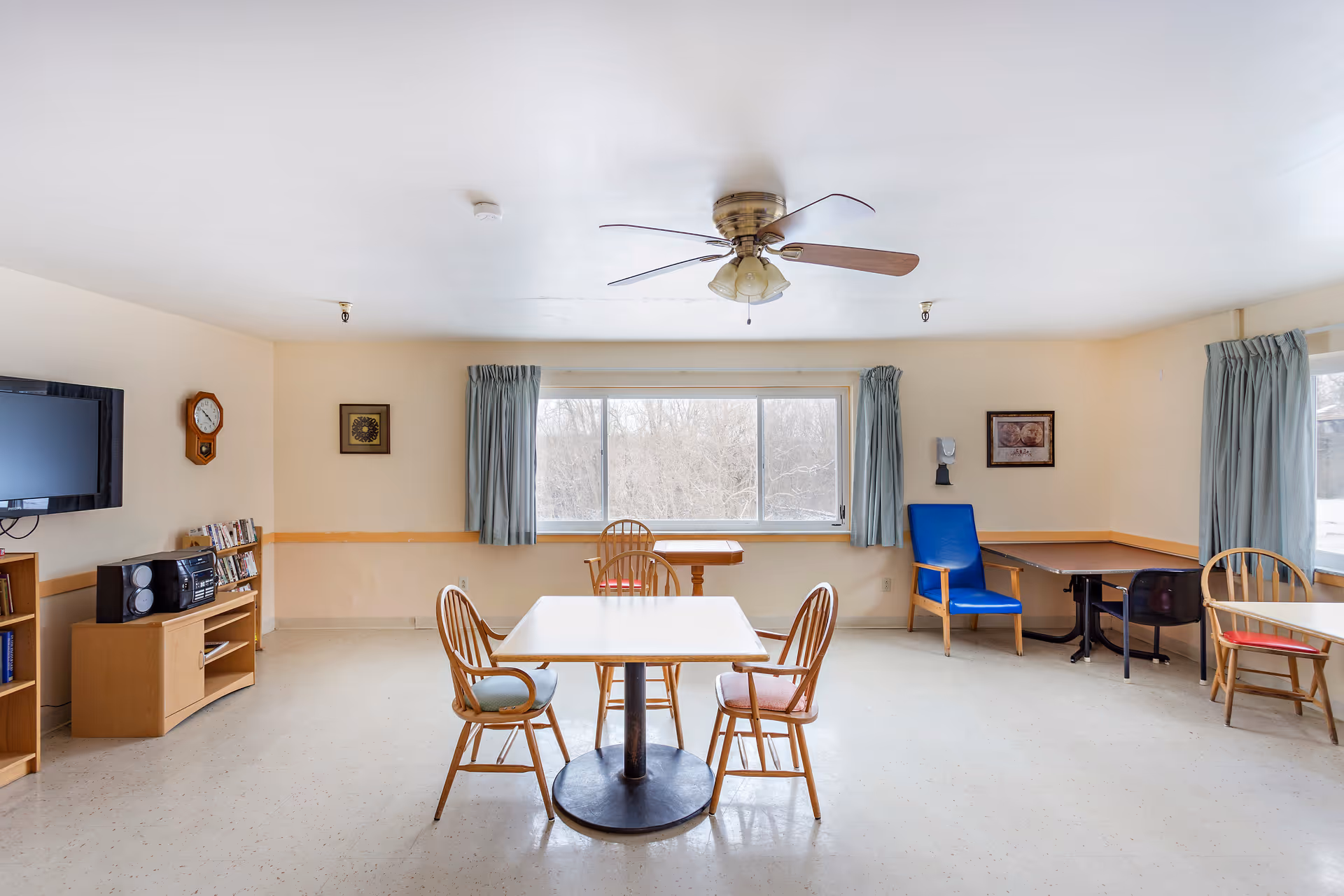 A bright, simple common room with a ceiling fan, several wooden tables and chairs with cushions, a blue armchair, a wall-mounted TV, a clock, a small bookshelf with books and a stereo, and large windows with light blue curtains letting in natural light.