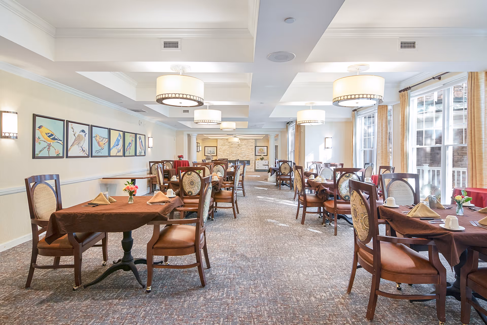A bright dining room with multiple tables covered in brown tablecloths, each set with folded napkins, cups, and small flower arrangements. The room features large windows with beige curtains allowing natural light to fill the space. The walls are decorated with framed pictures of birds, and the ceiling has modern round light fixtures.