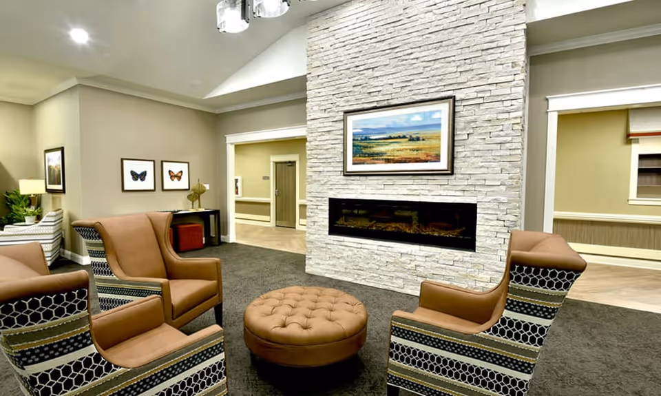 A cozy living room area with four patterned armchairs arranged around a round tufted ottoman. The room features a modern stone fireplace with a landscape painting above it. The walls are painted beige, and there are framed butterfly pictures on one wall. The space is well-lit with recessed ceiling lights.