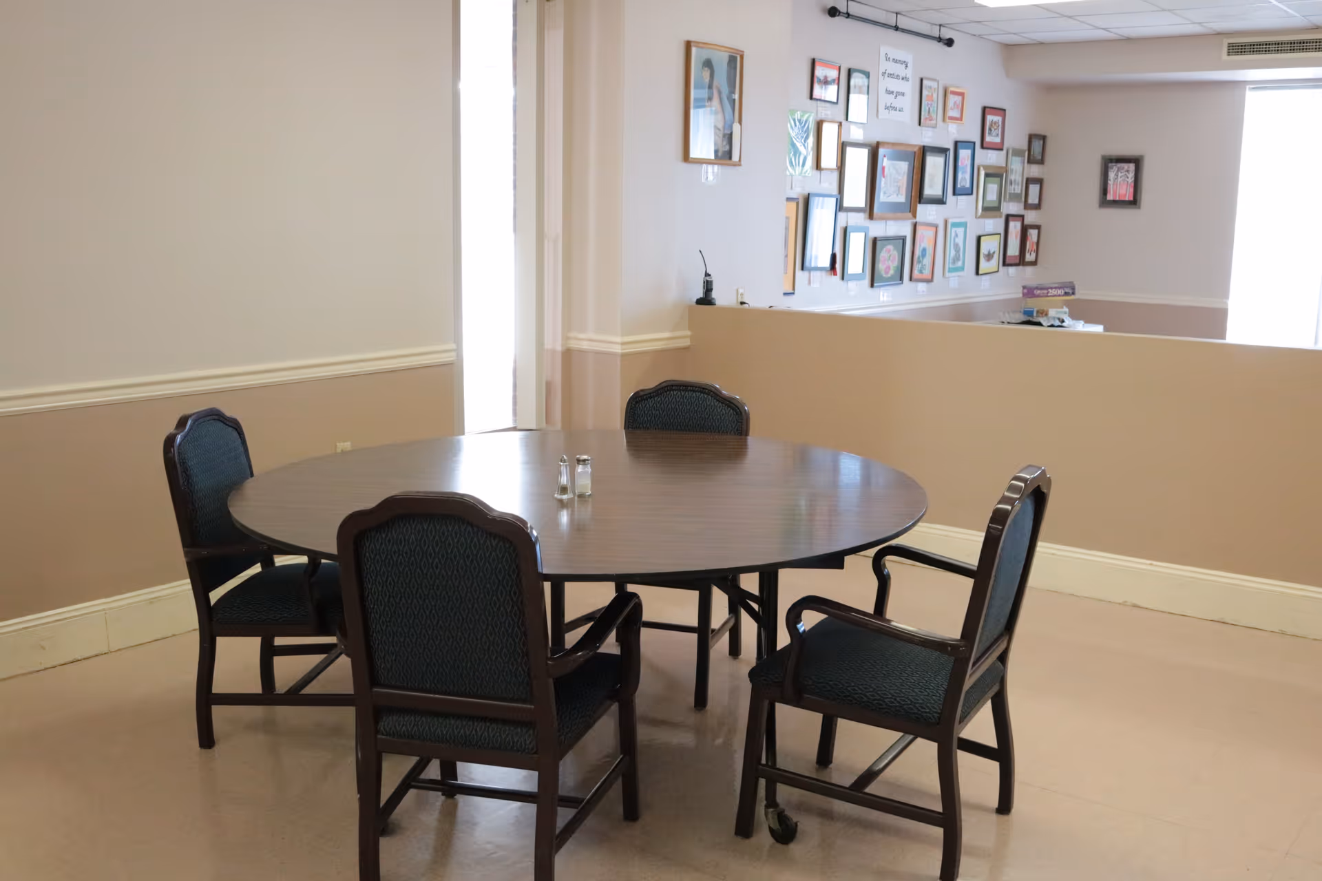 A round wooden table with four dark green cushioned chairs around it in a room with beige walls and a gallery wall of framed pictures and artwork in the background.