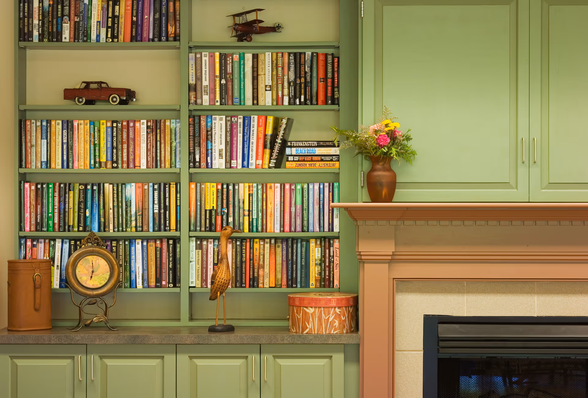Built-in green bookshelves filled with books and decorative items next to a fireplace mantel topped with a vase of flowers.