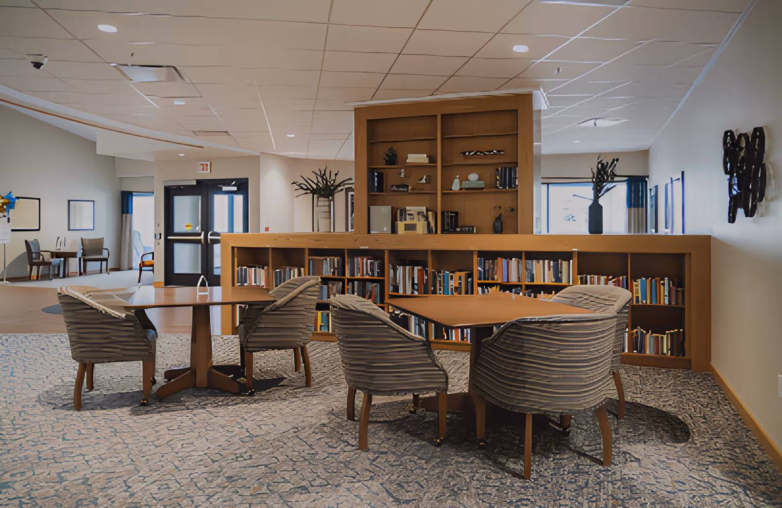 A cozy seating area in a senior living facility with two wooden tables surrounded by four striped upholstered chairs. Behind the seating area is a wooden bookshelf filled with books and decorative items. The room has a carpeted floor with a patterned design, light-colored walls, and a ceiling with recessed lighting. There are windows and doors in the background letting in natural light.