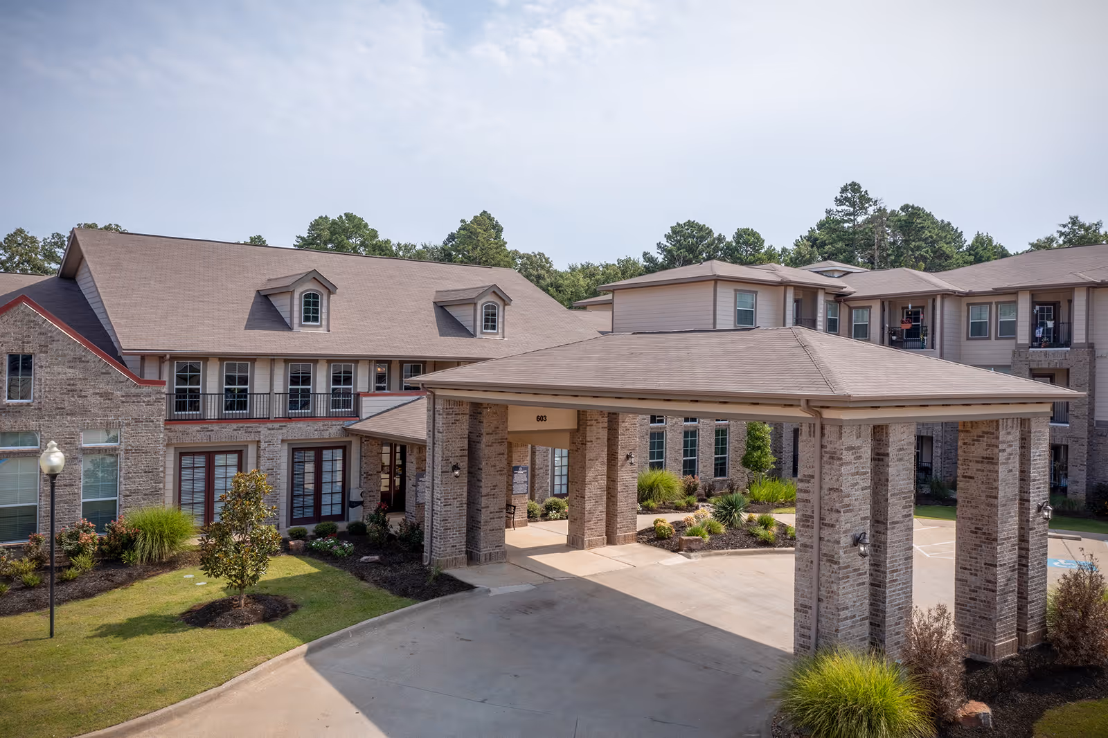 Exterior view of Parkview on Hollybrook senior living facility showing a large covered entrance with brick pillars, manicured landscaping, and a multi-story building with windows and balconies in the background under a partly cloudy sky.