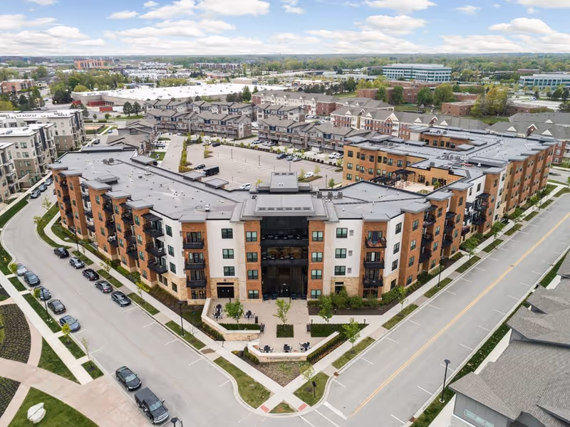 Aerial view of a large, modern senior living facility with multiple connected buildings, surrounded by roads and parking areas. The buildings have a mix of brick and light-colored exterior walls with balconies. The surrounding area includes other residential and commercial buildings under a partly cloudy sky.