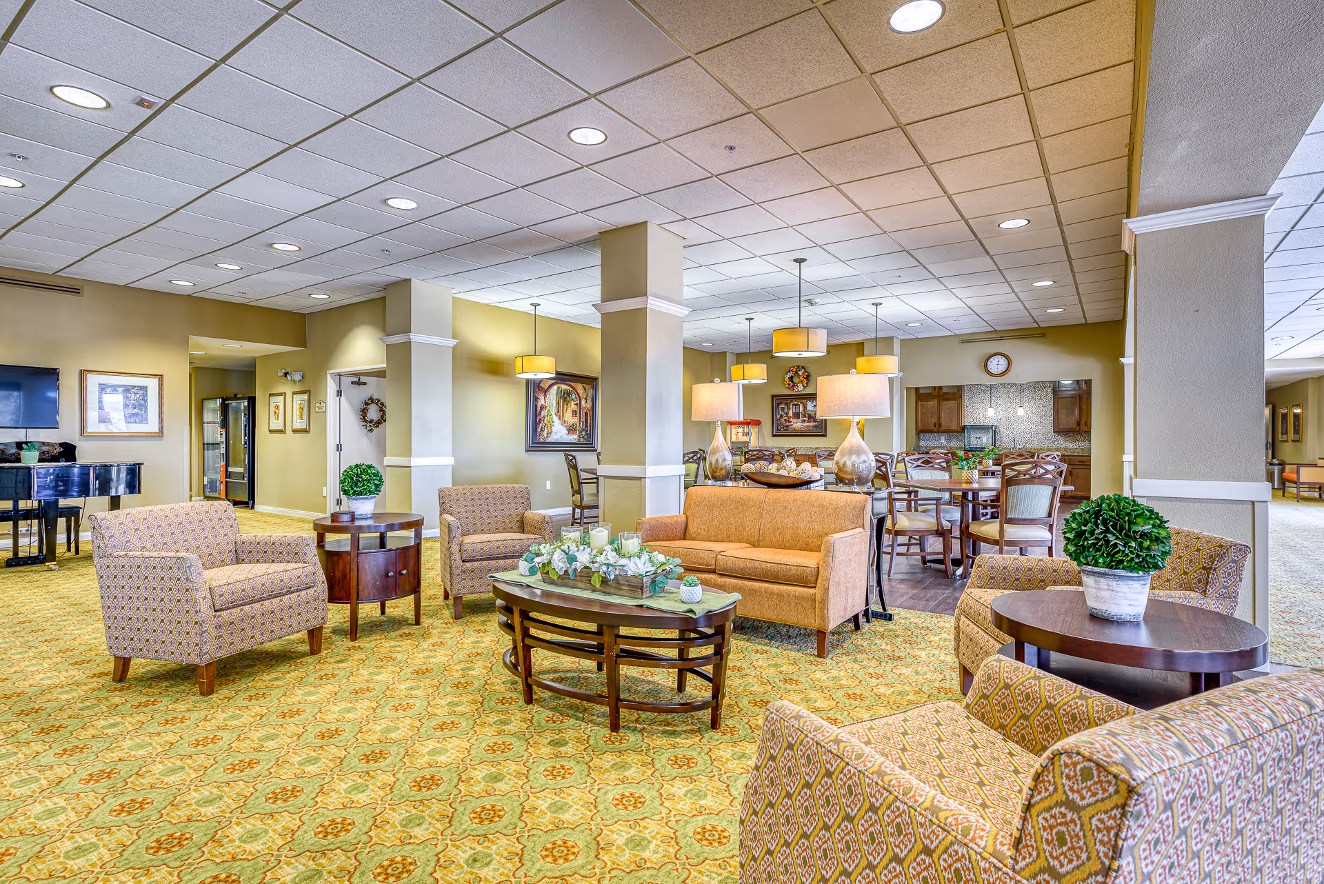 Spacious senior living common area with upholstered chairs and sofas, coffee tables, and a dining area in the background under warm lighting.