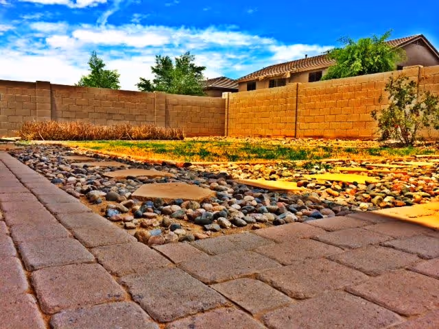 A backyard area with a paved stone walkway and a section filled with small rocks. The yard is enclosed by a tan brick wall, with some sparse grass and a few small plants. In the background, there are neighboring houses and a bright blue sky with some clouds.