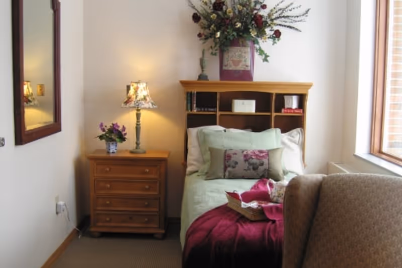 Sunlit single bedroom with a bed topped with pillows and a burgundy throw, a wooden headboard with shelves, a bedside drawer and lamp, and a window.