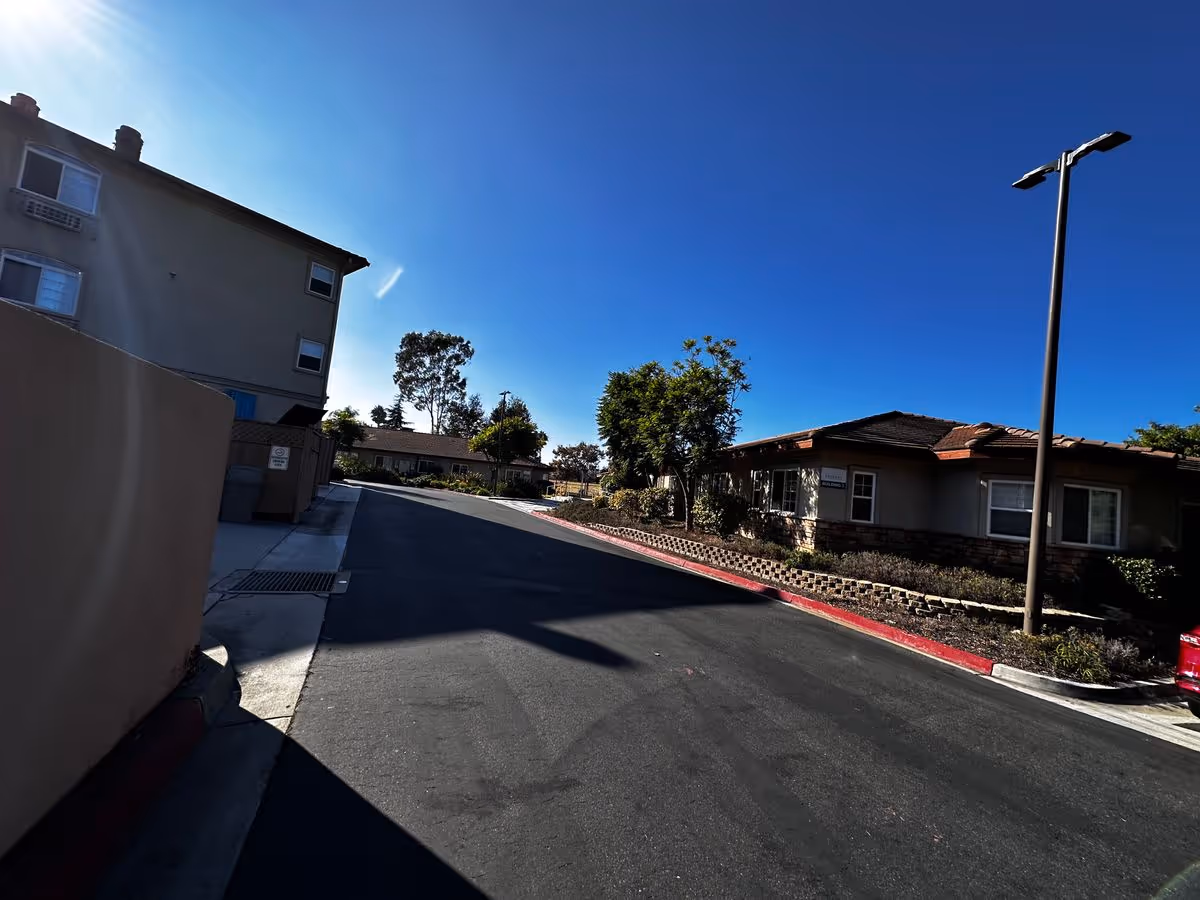 Wide exterior view of a senior living complex roadway lined with one-story buildings, landscaping, and a tall streetlamp under a clear blue sky.