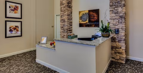 Reception area with a granite countertop desk, stone pillar accents, a potted plant, framed artwork on the walls, and a patterned carpeted floor.