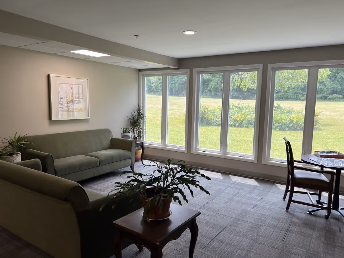A bright living room area with large windows overlooking a green outdoor space. The room features two green sofas, a wooden side table with a potted plant, another potted plant near the window, a framed artwork on the wall, and a small round table with two chairs near the windows.