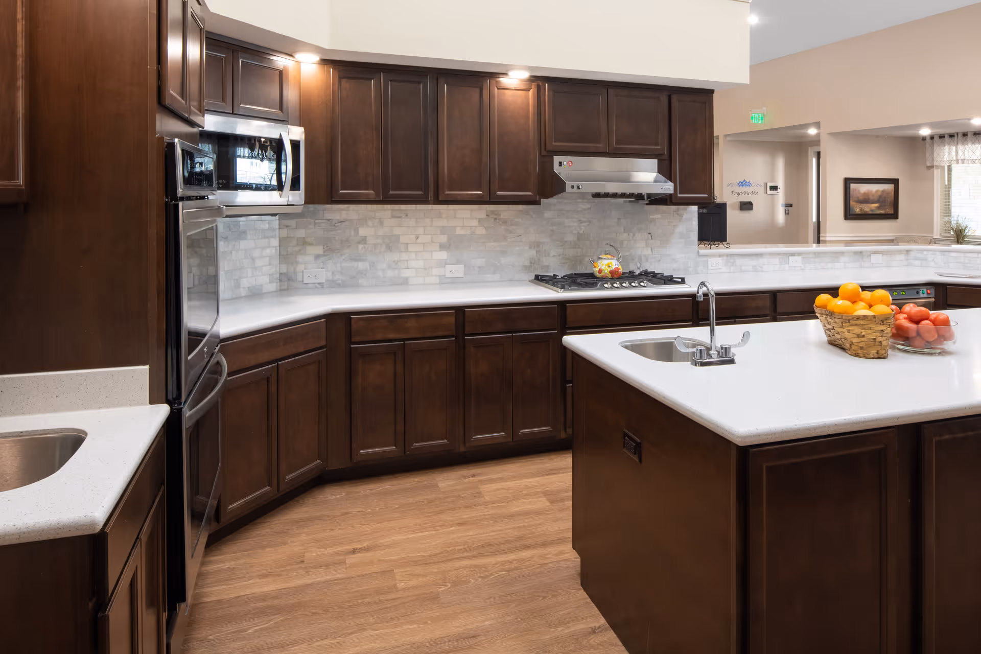 Modern kitchen with dark wood cabinets, white countertops, a built-in microwave and oven, a gas stove with a range hood, and a kitchen island with a sink. A basket of oranges and a bowl of tomatoes are on the island. The floor is wood, and there is a partial view into an adjacent room with a framed picture on the wall.