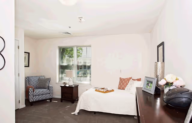 A bright and cozy bedroom with a single bed covered in a white quilt and decorative pillows. A wooden nightstand with a lamp is positioned next to a window with blinds, allowing natural light to enter. There is a patterned armchair in the corner and a wooden dresser with framed photos, a vase of flowers, and a radio on top. The walls are painted white, and the floor is carpeted in a neutral color.