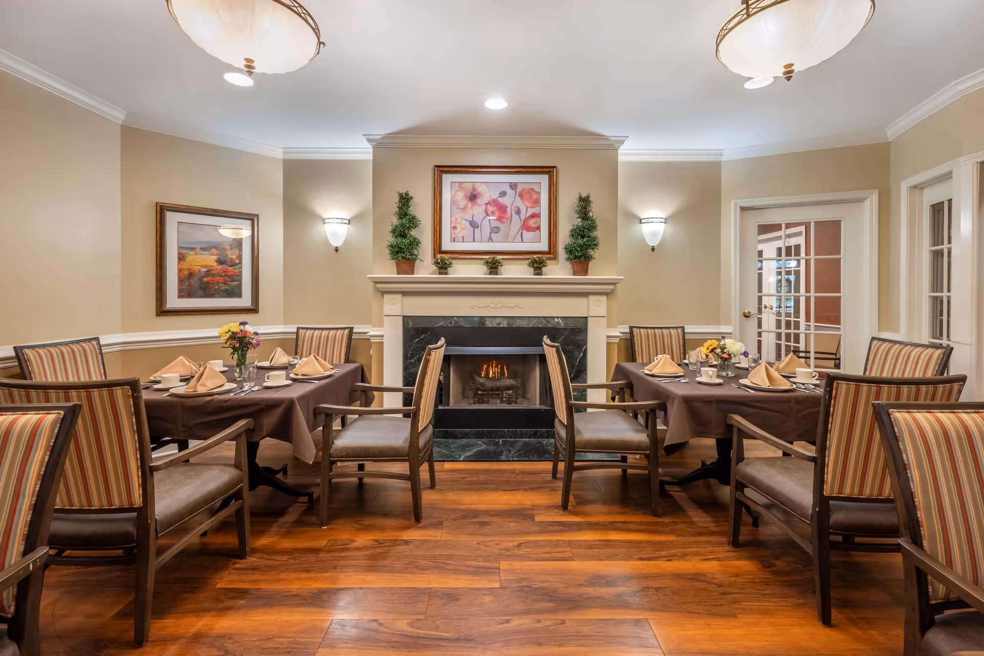 Formal dining room with tables set for a meal, striped chairs, hardwood floors, and a central fireplace topped with artwork.