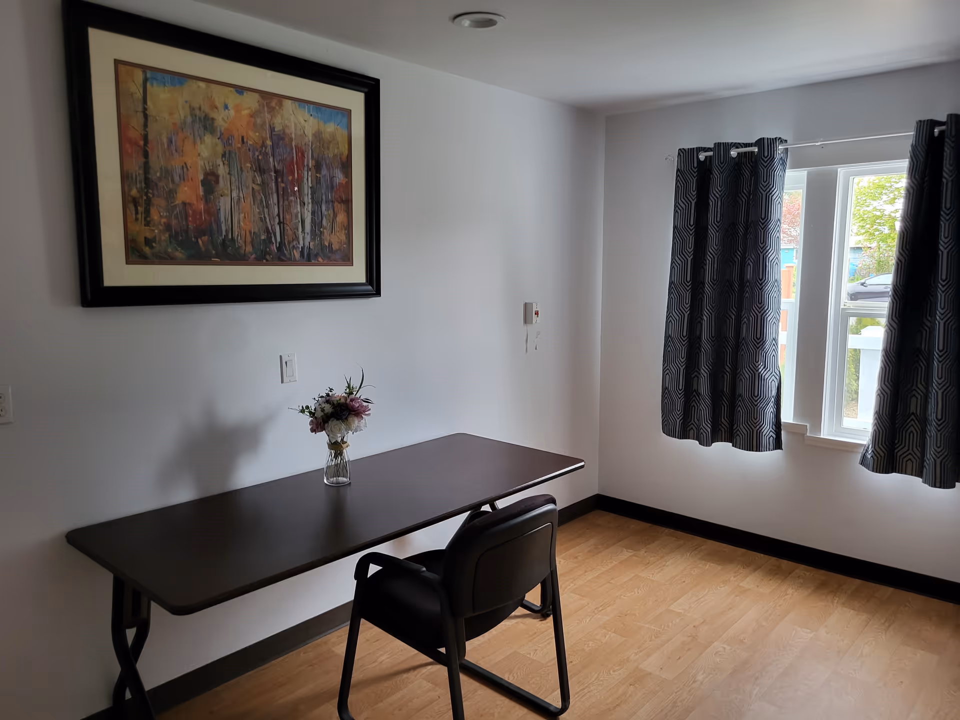A simple room with a dark rectangular table and a black chair. On the table is a small vase with a bouquet of flowers. A large framed painting of an autumn forest hangs on the white wall above the table. To the right, there is a window with patterned dark curtains partially open, letting in natural light. The floor is wooden.
