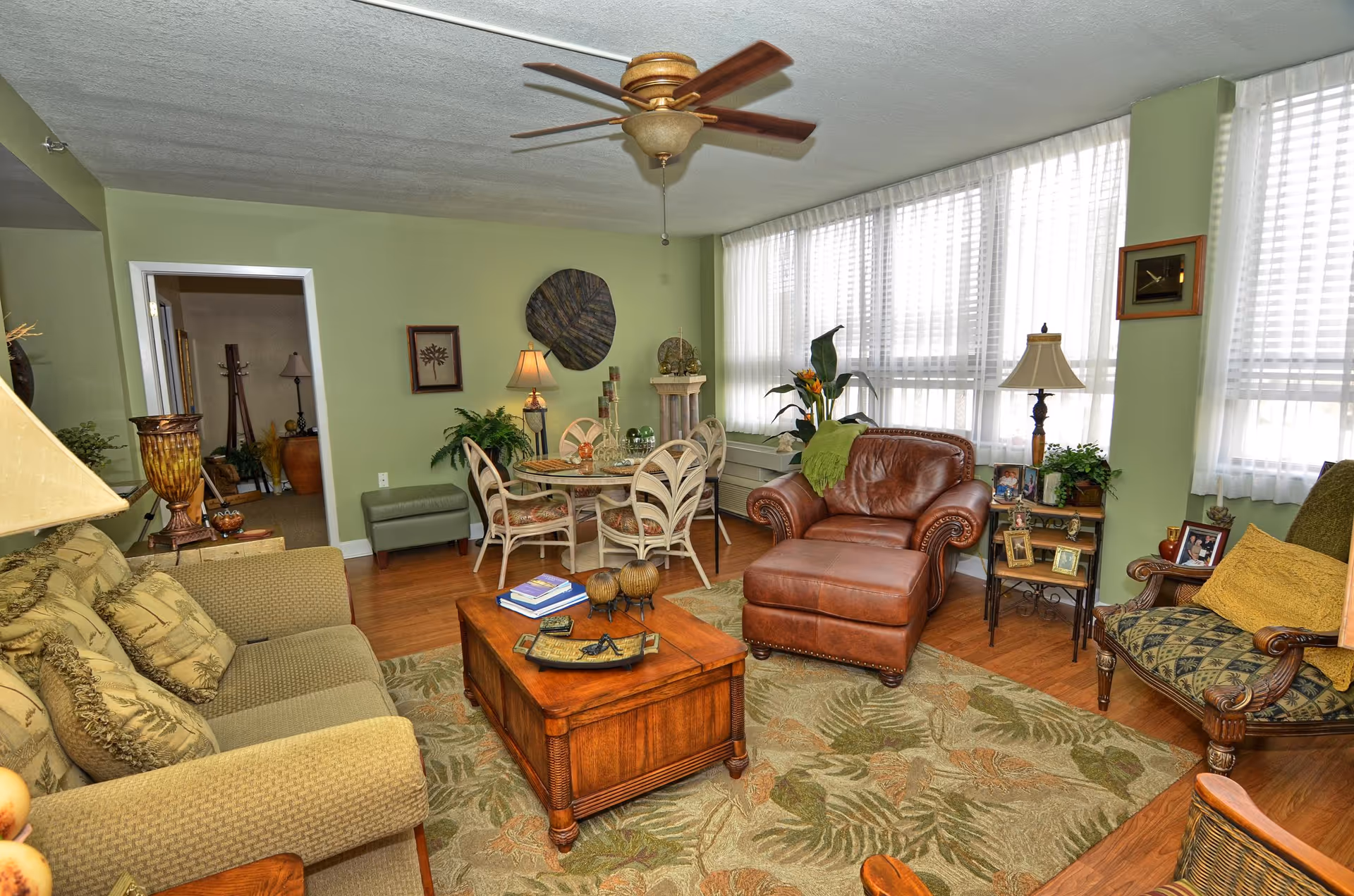 A cozy living room with green walls and large windows covered with sheer white curtains. The room features a green sofa with patterned cushions, a brown leather armchair with an ottoman, a wooden coffee table with decorative items and books, and a round glass dining table with six chairs. There are various plants, lamps, framed pictures, and wall decorations adding warmth to the space. A ceiling fan with wooden blades is mounted on the ceiling.