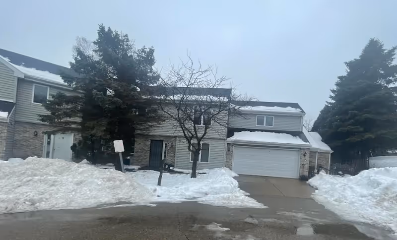 Exterior view of a residential-style building with snow-covered ground and driveway. The building has light-colored siding, a garage door, and a few windows. There are two large evergreen trees and one leafless tree in front of the building under a cloudy sky.