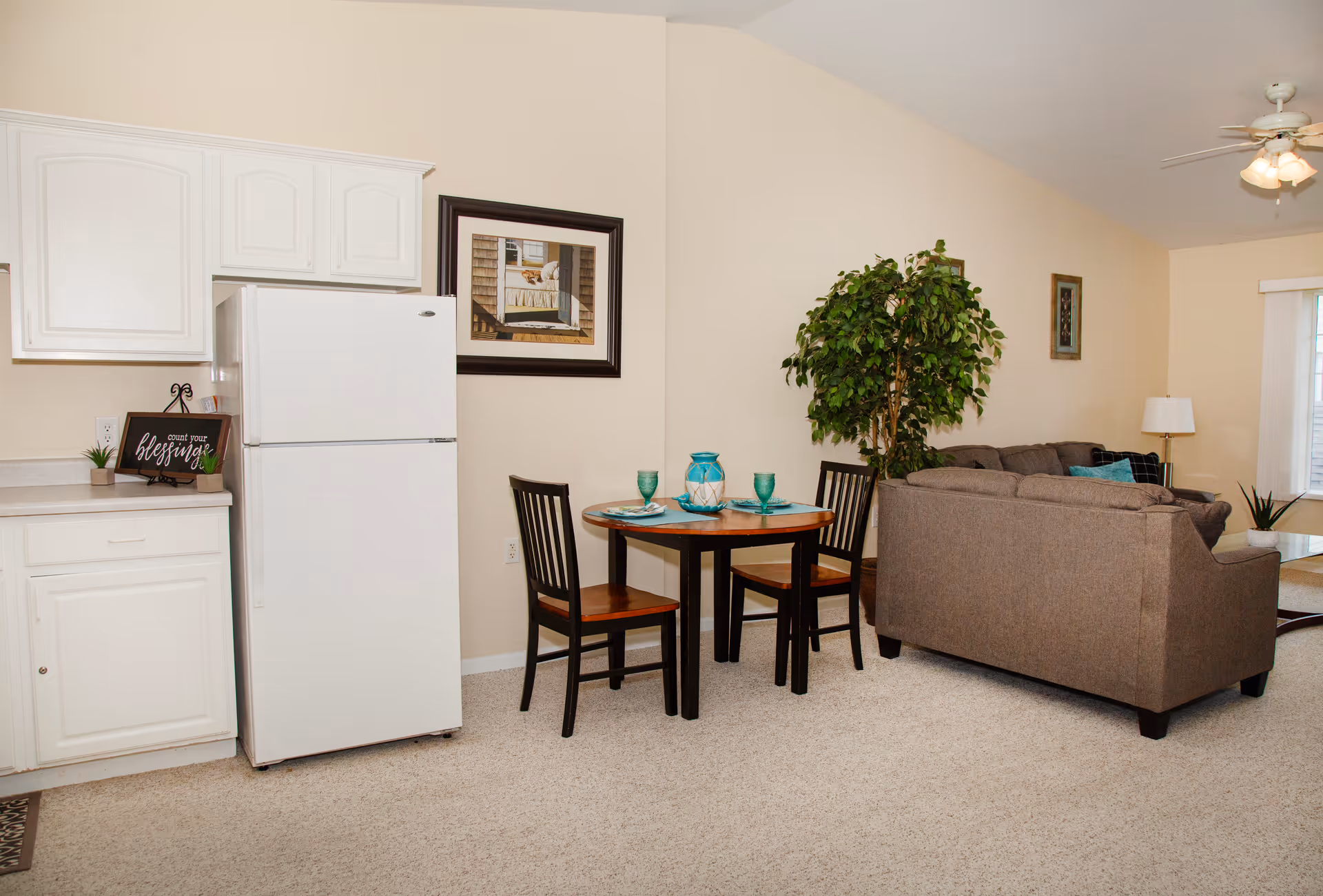 Interior view of a living space in an assisted living facility showing a small dining area with a round table set for two, a brown sofa, a large green potted plant, a ceiling fan with lights, and part of a kitchen with white cabinets and a white refrigerator.