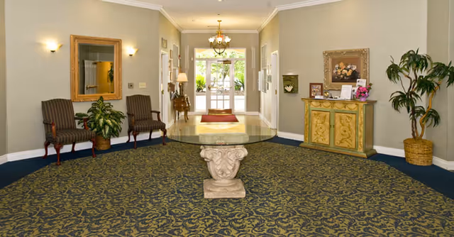 Furnished lobby with a round glass-top center table on patterned carpet, chairs and a console cabinet, and double doors leading outside.