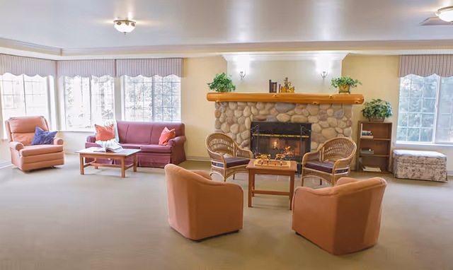 A cozy living room area in Birchview Memory Care featuring a stone fireplace with a wooden mantel, two wicker chairs with cushions, two upholstered armchairs, a maroon sofa, a coffee table with a book, and large windows letting in natural light. There are plants on the mantel and a small bookshelf to the right.