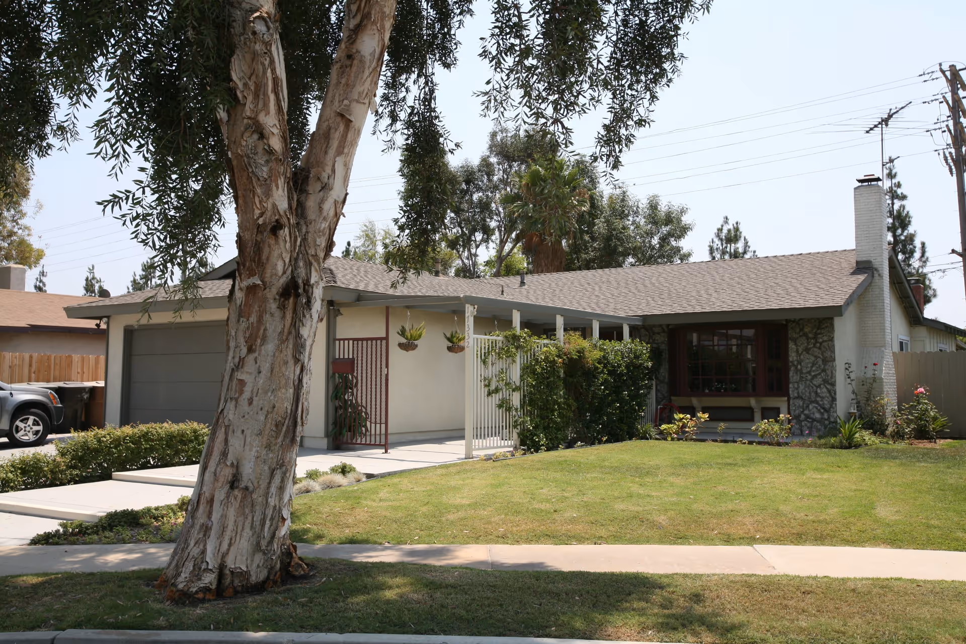 Front exterior view of a single-story house with a large tree in the foreground, a well-maintained lawn, and a driveway with a parked car. The house has a garage, a covered porch with hanging plants, and a chimney.