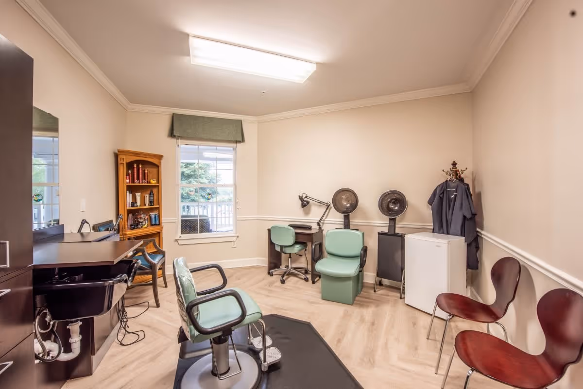 Interior view of a small salon or beauty room with a salon chair in the center, two hair dryer chairs against the wall, a small desk with a lamp, a wooden shelf with hair products, two maroon chairs, a coat rack with hanging garments, and a window with a green valance.