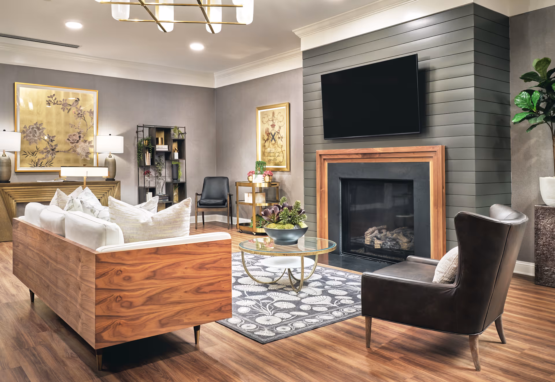 A cozy senior living room featuring a wooden sofa with white cushions, a dark leather armchair, a glass coffee table with a plant centerpiece, a fireplace with a wooden frame, and a wall-mounted TV above it. The room has wooden flooring, decorative paintings on the walls, a bookshelf, and a small table with plants and flowers.