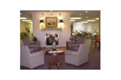 A cozy common area in a senior living facility with two upholstered armchairs facing each other around a small wooden table with a vase of flowers. The room has soft lighting, a fireplace with a framed picture above it, and additional seating and tables in the background.