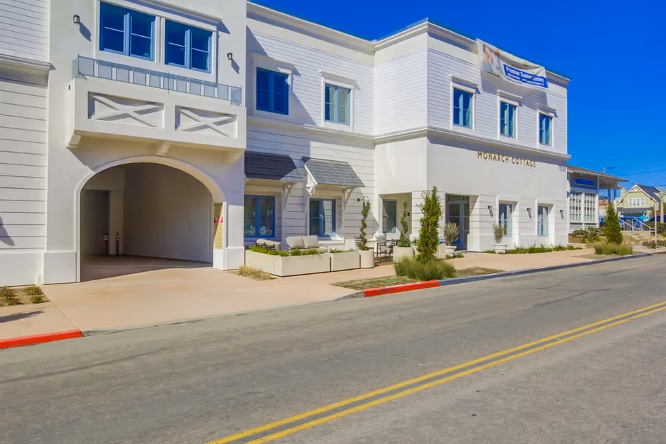 Exterior view of Monarch Cottages, a two-story white building with multiple windows, a covered driveway, and outdoor seating areas with plants along the sidewalk under a clear blue sky.