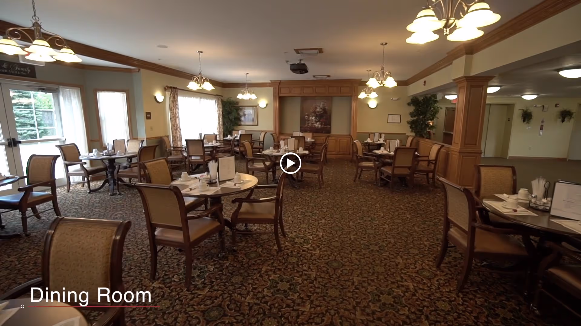 A spacious dining room with multiple round tables and chairs arranged neatly. Each table is set with white cups, saucers, and napkins. The room has patterned carpet flooring, beige walls with wooden paneling, and several light fixtures hanging from the ceiling. Large windows with curtains allow natural light to enter the room. There are plants and framed artwork on the walls, creating a warm and inviting atmosphere.