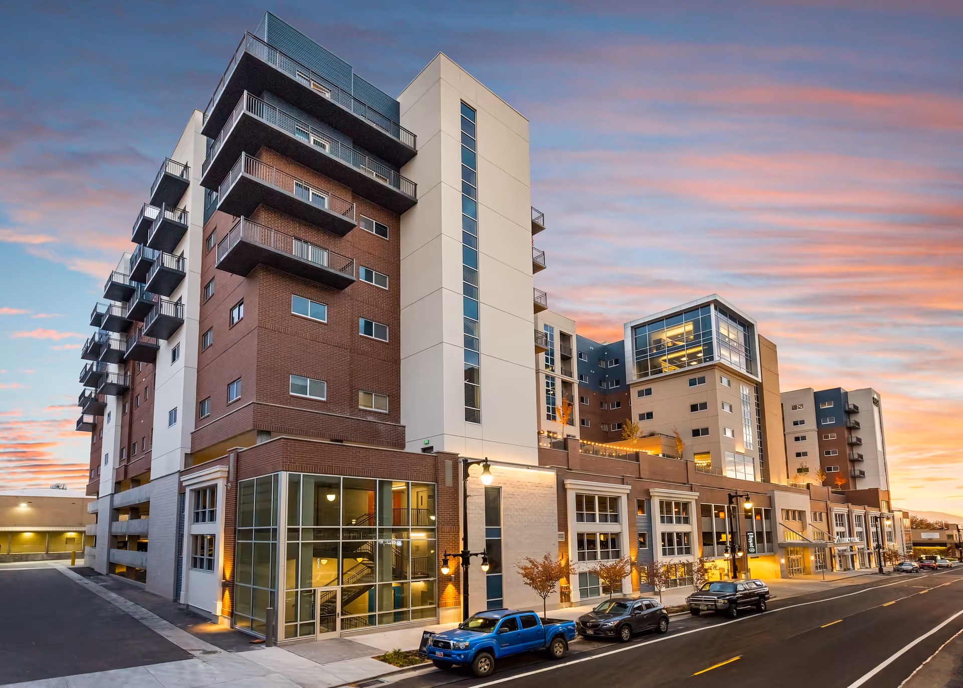 Exterior view of a modern multi-story senior living facility building at sunset with a partly cloudy sky. The building features large windows, balconies, and a mix of brick and light-colored exterior walls. Several cars are parked along the street in front of the building.
