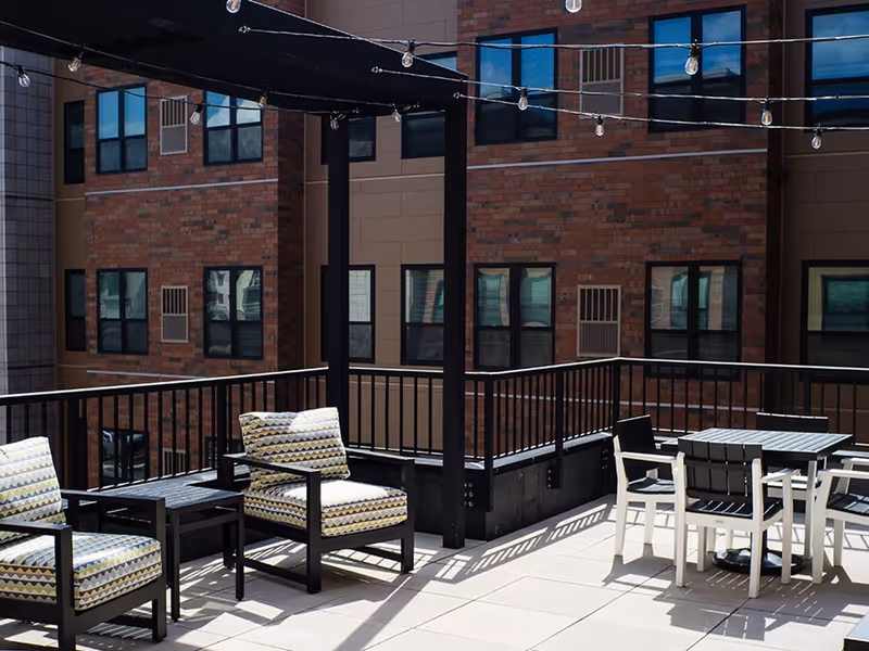Outdoor patio area with cushioned chairs, a small table, and a dining table with chairs, surrounded by a black metal railing and string lights overhead, with a brick building in the background.