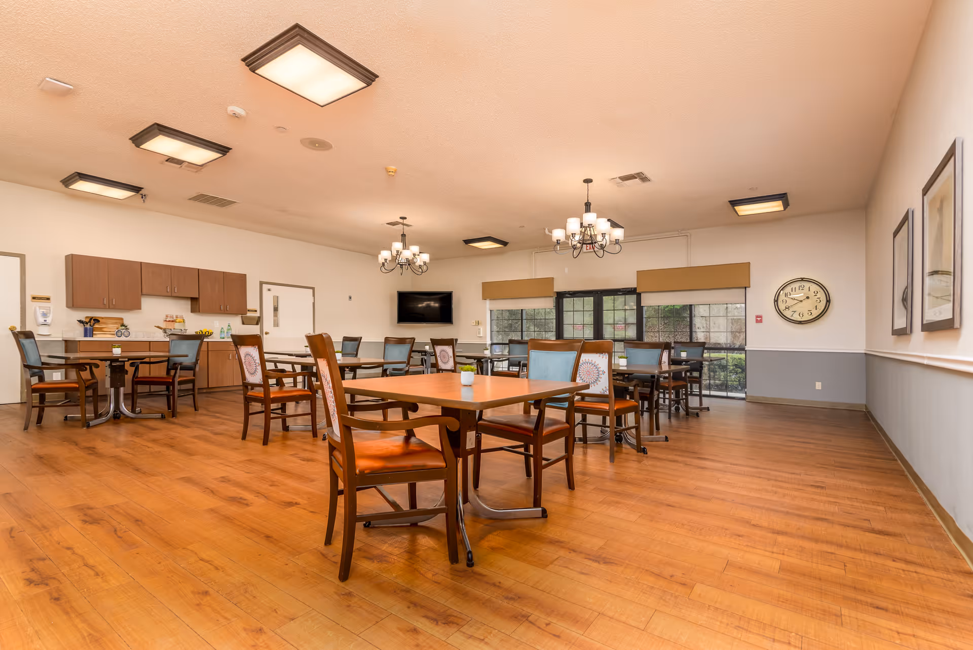 Spacious senior living dining/activity room with wooden tables and chairs, chandeliers, a wall clock, TV, and a small kitchenette area.