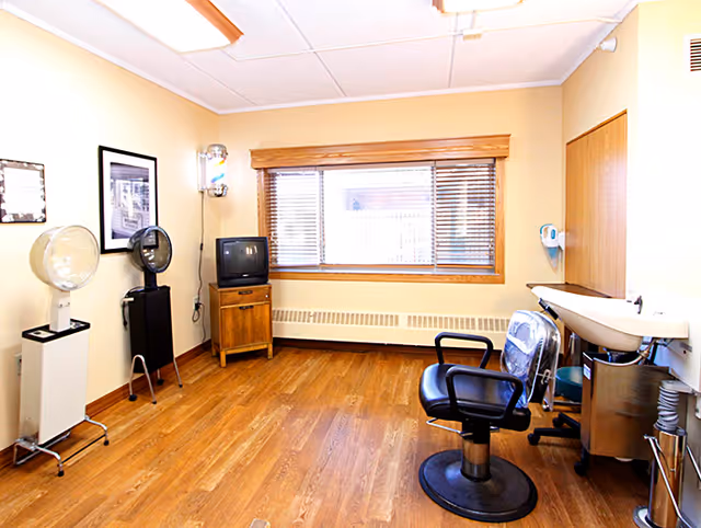 A small salon room with wooden flooring and beige walls. The room contains a black salon chair in front of a white sink with a faucet. There are two hair dryers on stands against the left wall, a small wooden cabinet with an old-style TV on top, and a large window with wooden blinds letting in natural light.