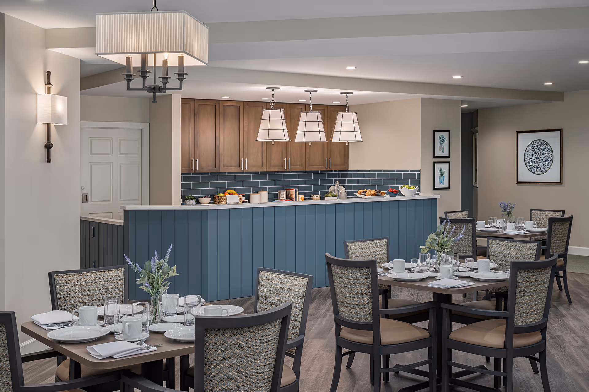 A dining area in Meredith Court featuring several tables set with white plates, cups, and napkins. Each table has a small floral centerpiece. The room has wooden chairs with patterned upholstery and a blue paneled counter with wooden cabinets and blue tile backsplash behind it. Three pendant lights hang above the counter, and framed artwork decorates the walls.