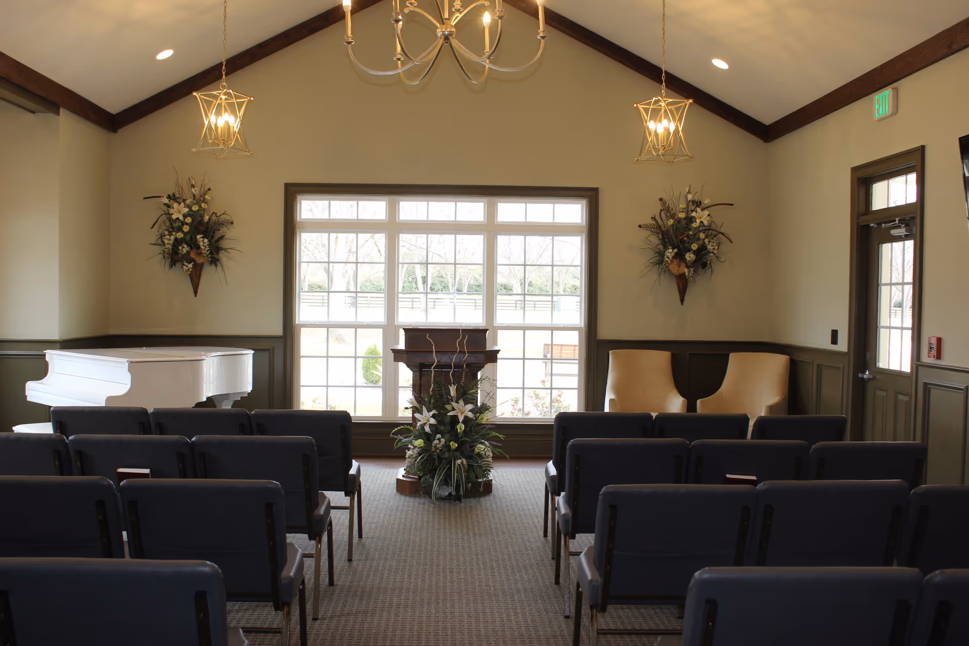 Interior room with rows of chairs facing a wooden podium adorned with a floral arrangement. Large windows behind the podium let in natural light. A white grand piano is on the left side, and two beige armchairs are on the right. The room has high ceilings with exposed wooden beams and hanging light fixtures.