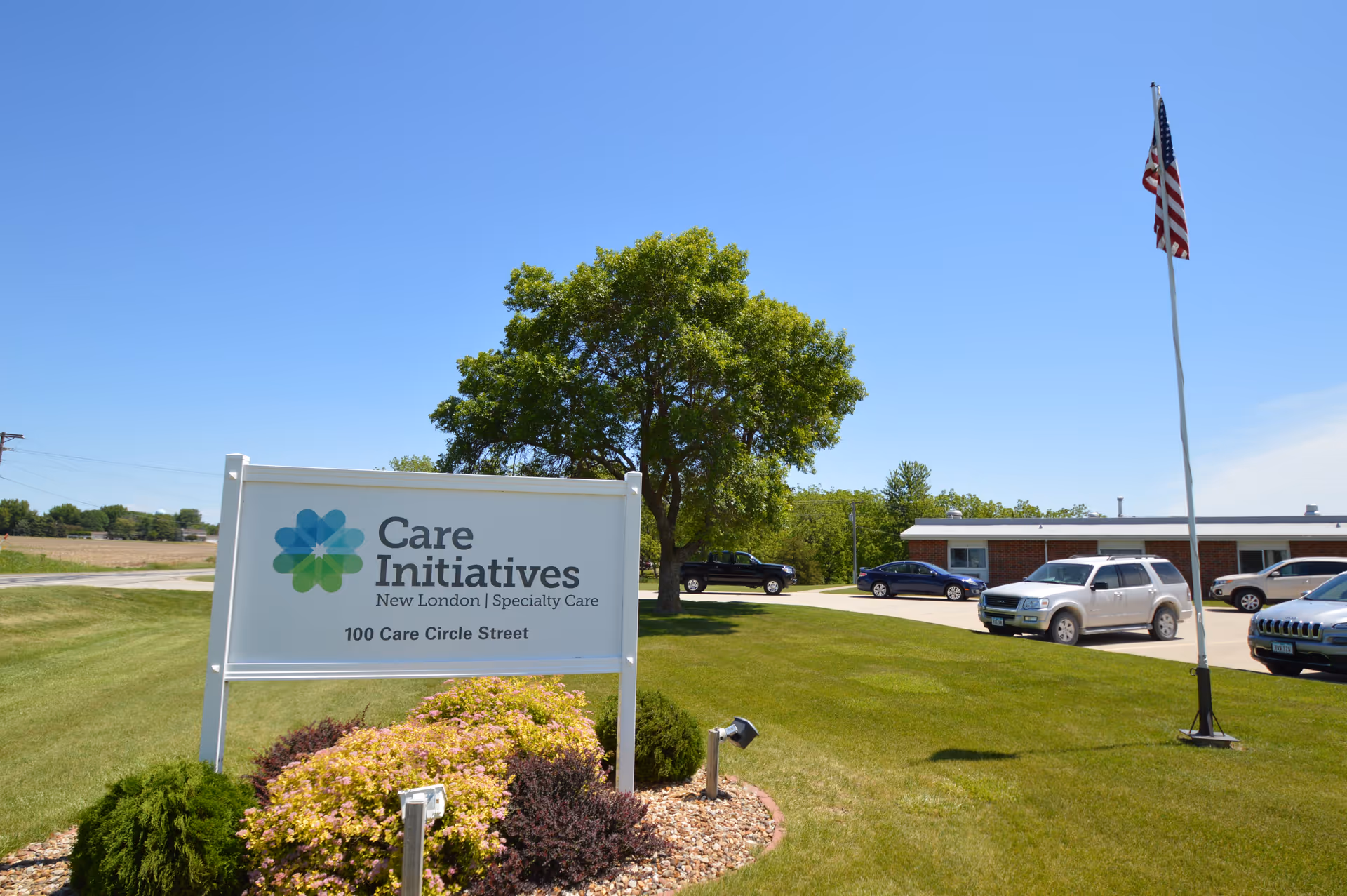 Outdoor view of Care Initiatives New London Specialty Care facility with a sign displaying the name and address 100 Care Circle Street, a green lawn, a tree, parked cars, and an American flag on a flagpole under a clear blue sky.