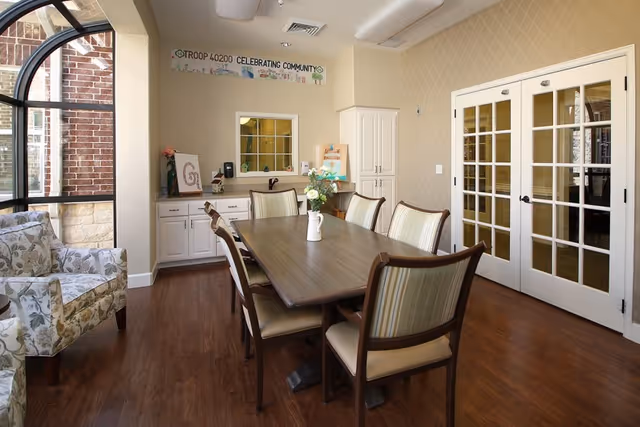 A bright dining room with a rectangular wooden table surrounded by six cushioned chairs. A vase with flowers is placed in the center of the table. To the left, there is a floral-patterned armchair near large windows with black frames, letting in natural light. The back wall features a small counter with cabinets, a window, and a banner that reads 'TROOP 40200 CELEBRATING COMMUNITY'. On the right side, there are white double French doors with glass panes. The room has wooden flooring and beige patterned wallpaper.