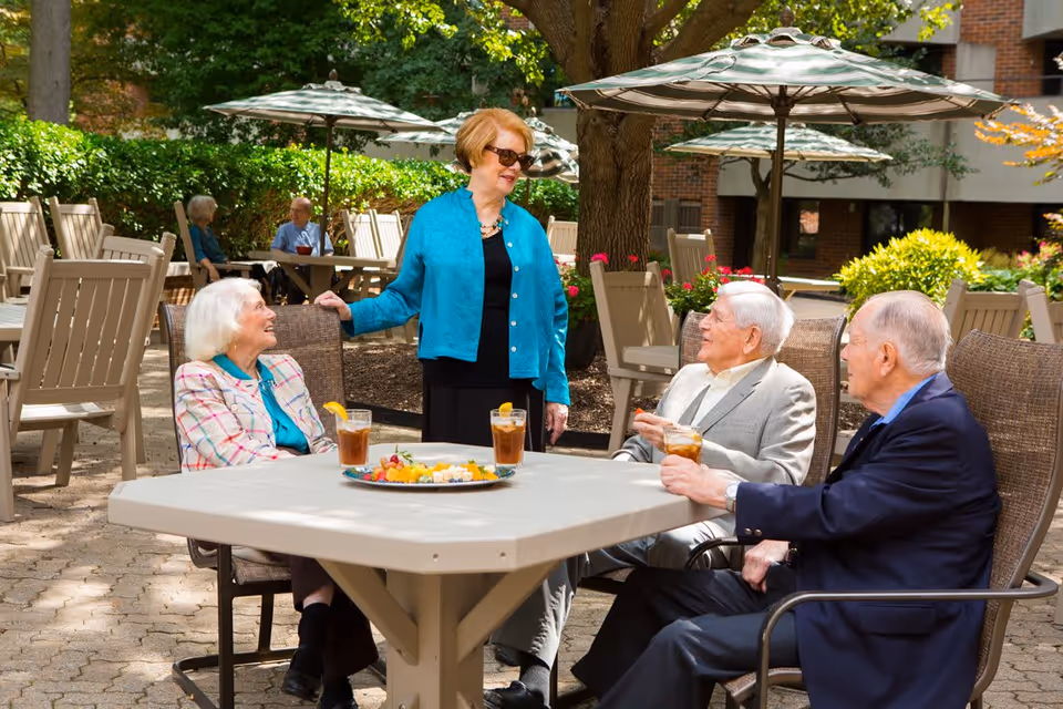 Four elderly people enjoying a sunny day outdoors at a senior living facility. Three are seated around a table with drinks and a plate of fruit, while one woman stands and smiles at them. The area is shaded by large umbrellas and surrounded by greenery and brick buildings.