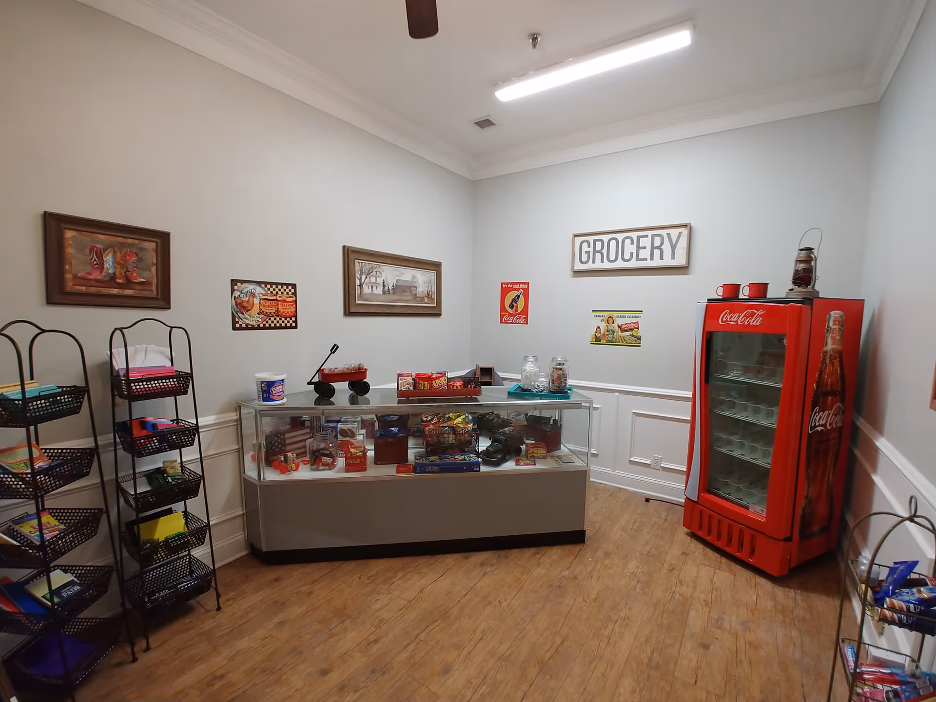 Interior view of a small grocery room with wooden flooring and light gray walls. There is a glass display case filled with snacks and small items in the center, a red Coca-Cola branded refrigerator on the right, and two black metal racks with various colorful items on the left. The walls are decorated with vintage-style signs and paintings, including a large sign that says 'GROCERY'.