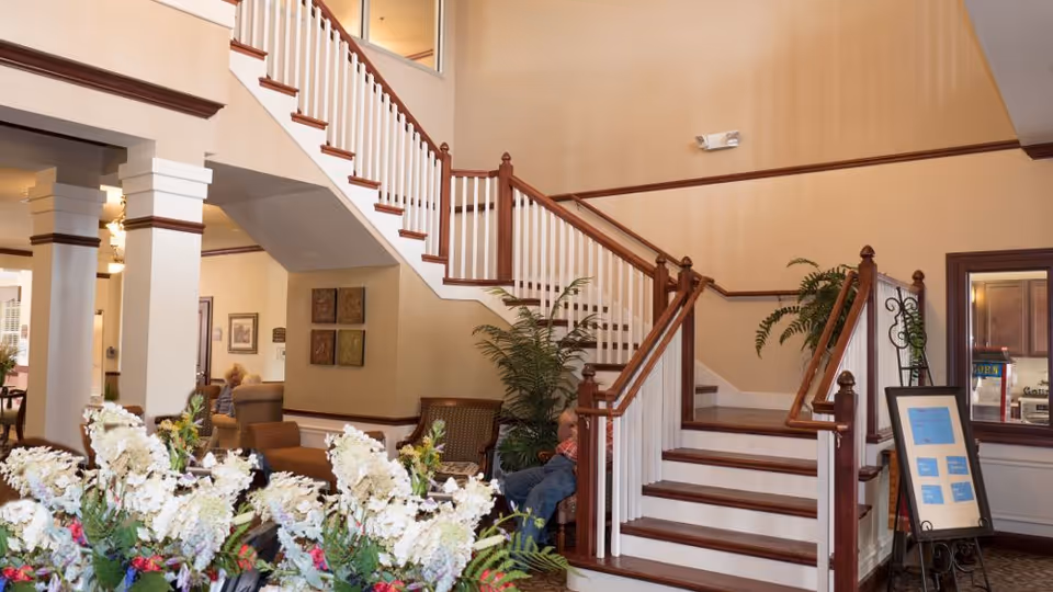 Interior view of a senior living facility lobby area with a staircase featuring white railings and wooden handrails. There are chairs and plants near the staircase, a floral arrangement in the foreground, and a person seated on a chair. The walls are painted beige with wooden trim, and there is a framed sign on an easel near the stairs.