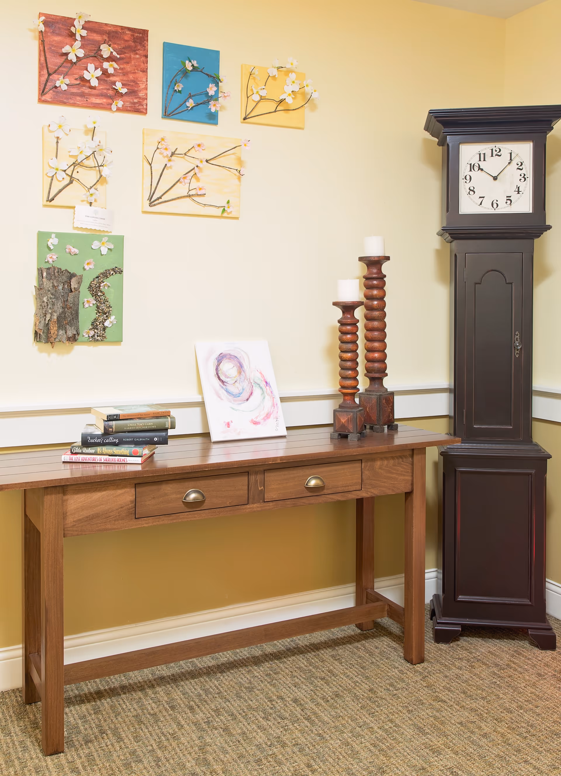 A wooden console table with two drawers against a beige wall. On the table are several stacked books, a small abstract painting, and two tall wooden candle holders with white candles. To the right of the table is a tall dark wooden grandfather clock showing the time as 10:10. Above the table on the wall are six decorative art pieces featuring branches with white flowers on colorful backgrounds.