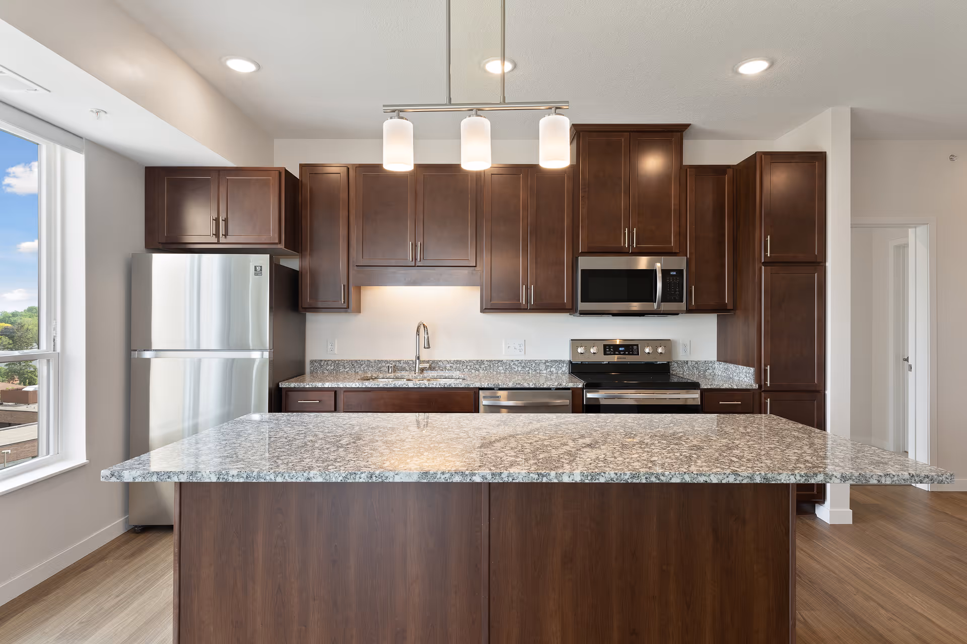 Modern kitchen with a large granite island, dark wood cabinets, stainless steel appliances and pendant lights.