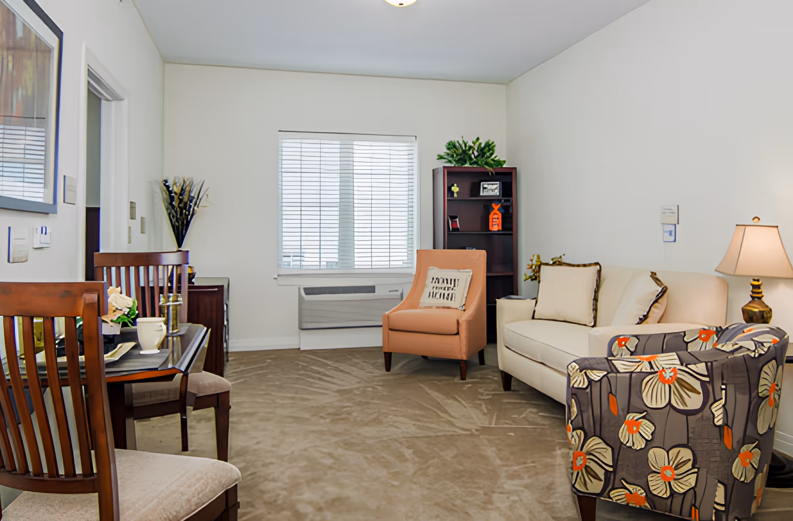 A cozy living room with a beige sofa, an orange armchair with a decorative pillow, and a floral-patterned armchair. There is a wooden bookshelf with plants and decorative items, a table lamp on a side table, and a window with white blinds letting in natural light. Two wooden chairs and a small table with a cup and flowers are visible on the left side.