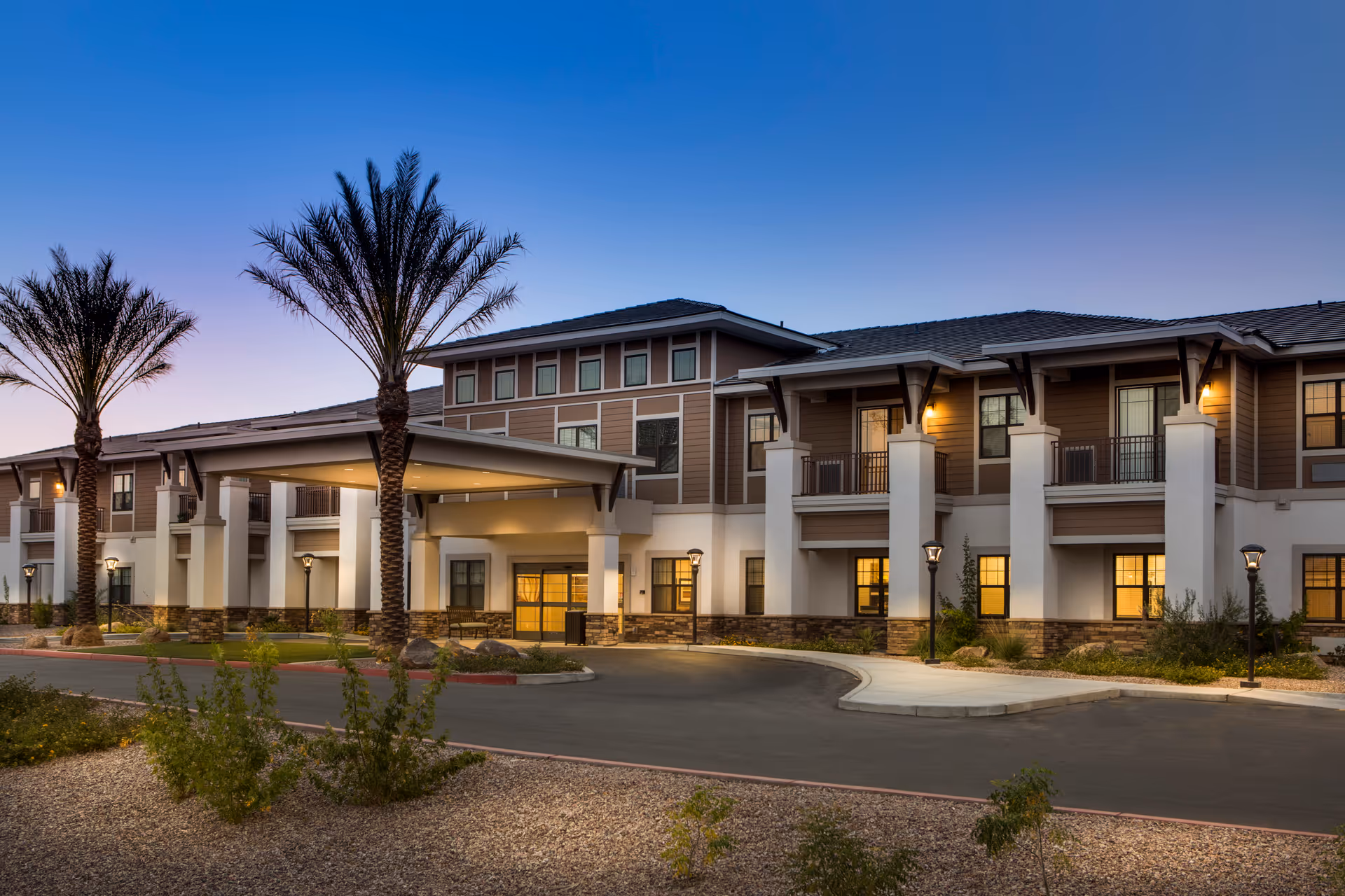 Exterior view of Savanna House senior living facility at dusk, showing a two-story building with a covered entrance, palm trees, and landscaped grounds with shrubs and gravel.
